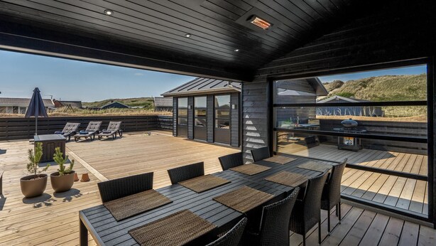 Un espace extérieur abrité doté d’une terrasse en bois, de sièges et de grandes fenêtres offrant une vue sur la plage et les dunes.