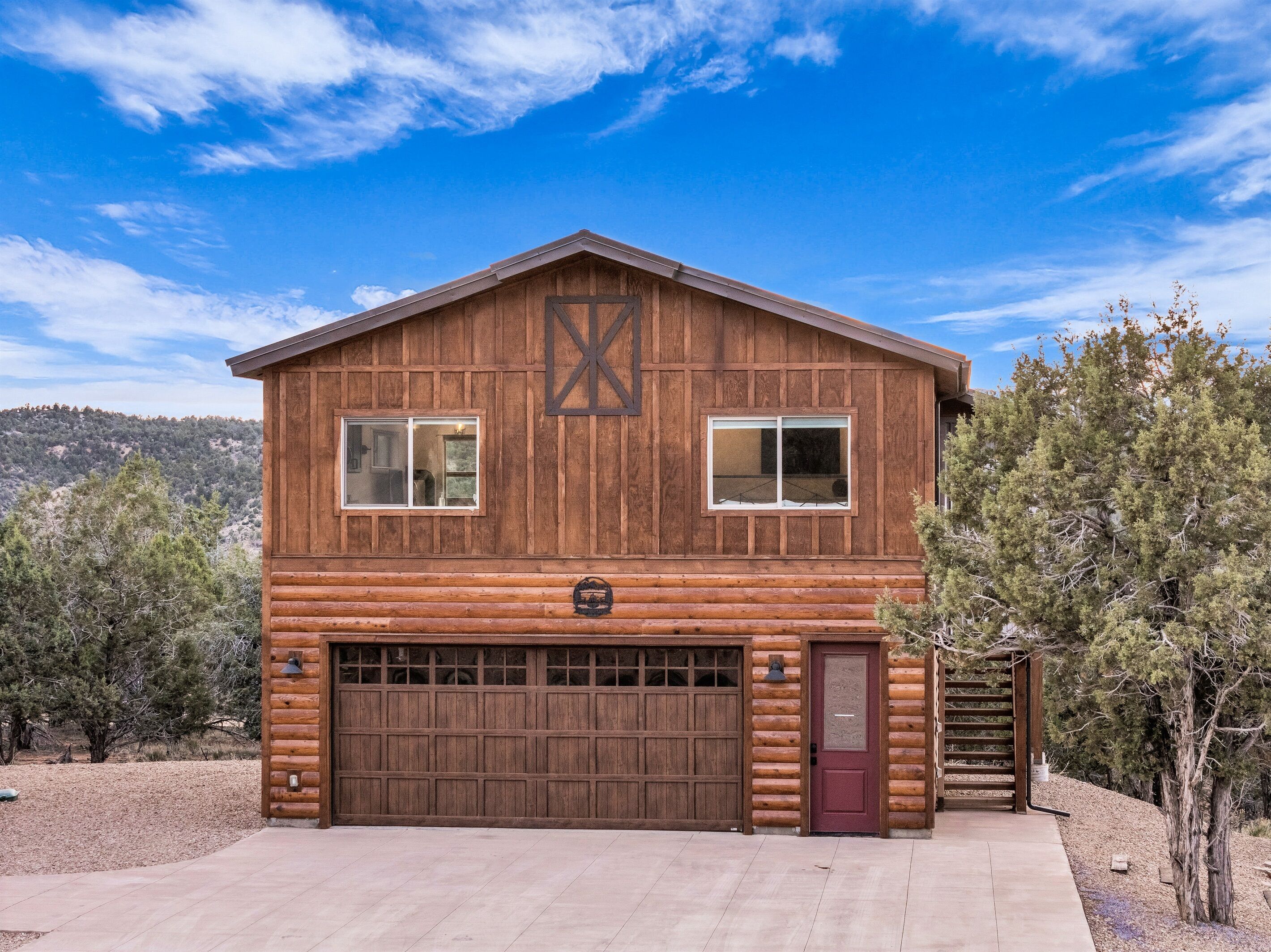A two-story log cabin with a large garage, a red door, and a wooden X-shaped design on the front wall.