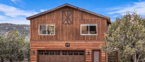 A two-story log cabin with a large garage, a red door, and a wooden X-shaped design on the front wall.