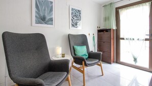 To grå lænestole med træben, et lille hvidt bord med lampe og et træskab med grøn gardin.