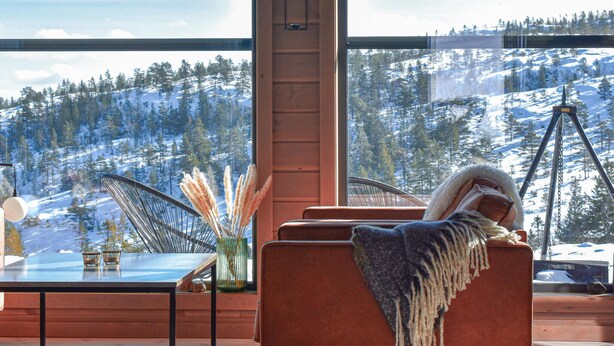 Ein gemütliches Zimmer mit großem Fenster und Blick auf verschneite Berge, ein bequemer Sessel mit warmer Decke sowie ein Tisch mit Pampasgras in einer Vase.