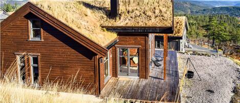 A wooden cabin with a grass roof, a wooden deck, and a view of the surrounding hills.