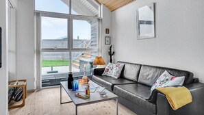 A modern living room with a leather sofa, a coffee table with various items, and a large window offering a view of a wooden deck and a house. - 2 bedroom beautiful home in Kerteminde (Kerteminde)