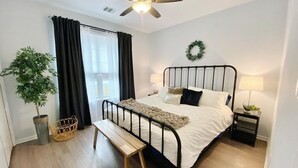 Une chambre à coucher comprenant un lit, des tables de chevet, un banc, un ventilateur de plafond, une fenêtre avec des rideaux, une plante en pot et une guirlande décorative accrochée au mur.