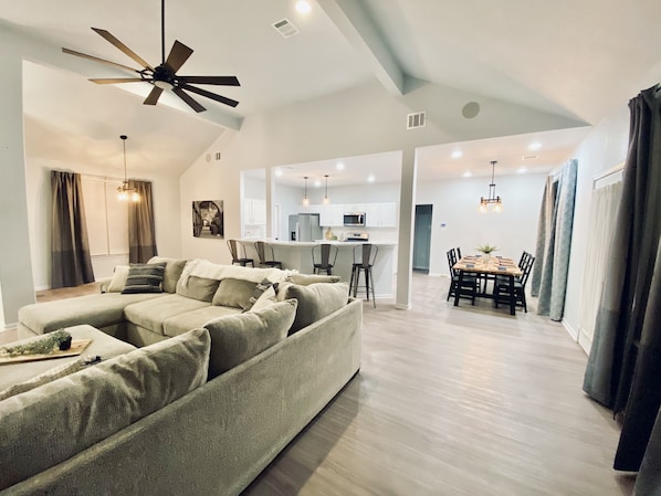 Relax in this open-plan living space with a large L-shaped sofa, dining area, and kitchen, featuring a high ceiling and exposed beams.