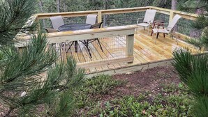 A wooden deck with a table and chairs, surrounded by a metal railing, set in a forested area.