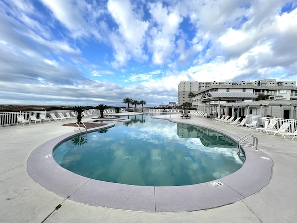 A large, circular swimming pool with lounge chairs and palm trees, surrounded by a white fence and buildings in the background.