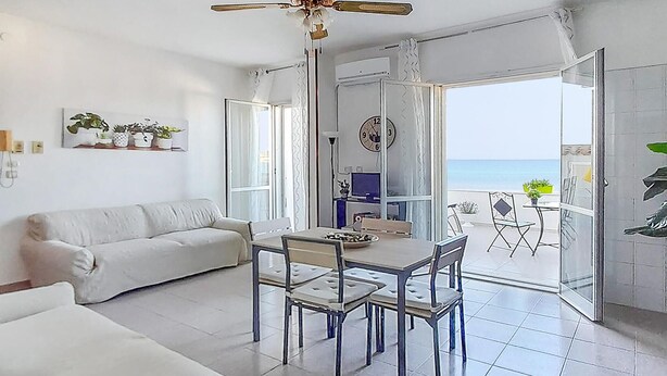 A modern living room with a white sofa, a dining table with chairs, and a view of the sea through an open door. - 1 bedroom beach front home in Siracusa (Siracusa)