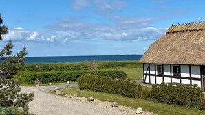 A thatched-roof house with a gravel path leading to it, surrounded by greenery and a view of the sea.