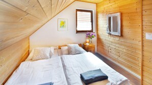 Un dormitorio con techo inclinado de madera, una cama con ropa de cama blanca, dos almohadas, una mesita de noche con una lámpara y una ventana con persianas.