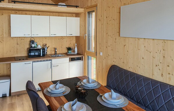 A compact kitchen with white cabinets, a microwave, and a sink. There is a dining table set with plates and a black cushioned bench. - Ferienhaus am Naturbadesee (Königsdorf)