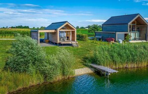 Two modern cabins with wooden exteriors, a covered outdoor seating area, and a dock extending into a calm lake. - Ferienhaus am Naturbadesee (Königsdorf)