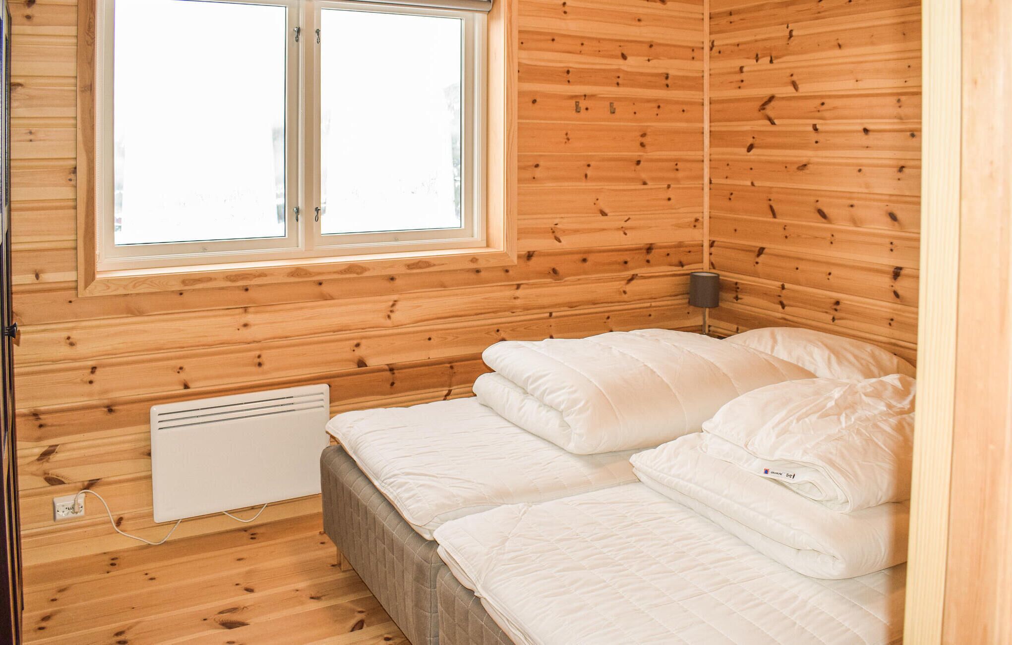 Une petite pièce chaleureuse aux murs en bois et dotée d’une seule fenêtre. On y trouve un lit avec une literie blanche et un radiateur posé sur le sol.