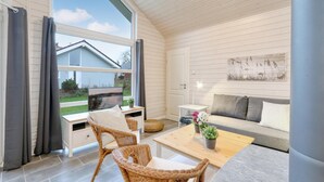 Ein gemütliches Zimmer mit einer Couch, einem Tisch und einem Korbstuhl. Ein Fenster mit Vorhängen, ein Bild an der Wand und Blick auf ein Haus.