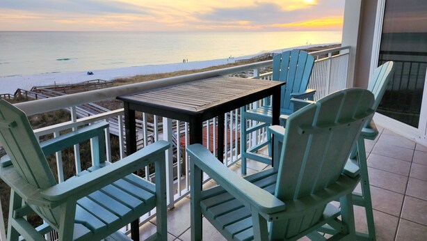 A balcony with a view of the ocean and a sunset. There are two chairs and a table. - Beachfront with Beach Setup Included! (Navarre)