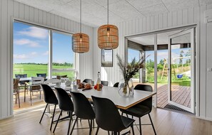 Un comedor con una mesa preparada para cuatro personas, dos lámparas de mimbre colgantes y vista a un campo verde a través de amplios ventanales.
