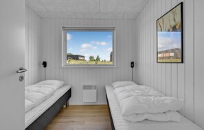 Un dormitorio pequeño con paredes blancas, dos camas individuales, una ventana con vistas a edificios y zonas verdes, y un cuadro en la pared.