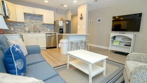 Un soggiorno moderno con un divano grigio, un tavolino da caffè bianco e una poltrona con motivi decorativi. Sullo sfondo, si intravede una cucina con mobili bianchi e una parete rivestita di piastrelle.