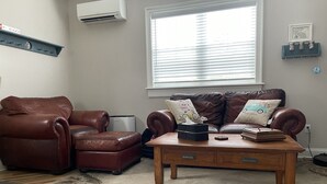 Ein Wohnzimmer mit braunem Ledersofa, einem Holztisch, einem gemusterten Teppich und einem Fenster mit Jalousien.