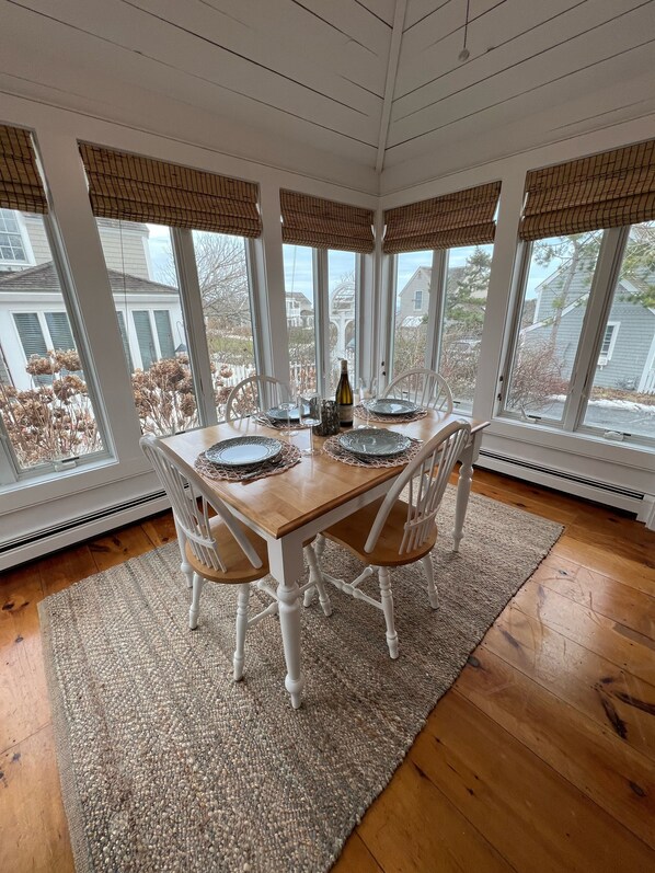 Un coin repas avec une table en bois dressée pour quatre personnes, entourée de chaises blanches, et offrant une vue sur les maisons à travers de grandes fenêtres.