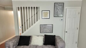 A living room with a grey tufted sofa, two black cushions, a white fluffy throw, and three framed artworks on the wall. - Spacious Atl Gem near the Battery & Six Flags! (Mableton)
