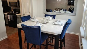A dining area with a white table set for four, blue upholstered chairs, a large mirror above the table, and a kitchen in the background. - Spacious Atl Gem near the Battery & Six Flags! (Mableton)