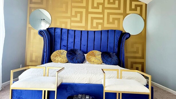 A bedroom with a blue upholstered headboard, a geometric patterned wall, two bedside tables with white cushions, and two round mirrors. - Spacious Atl Gem near the Battery & Six Flags! (Mableton)