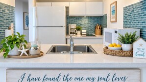 A kitchen with a white refrigerator, a sink, and a countertop with a decorative sign. - Beachfront  King Bed  Walk Everywhere (Carolina)