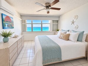 Un dormitorio con cama, ventilador de techo, ventana con vista al océano, cómoda y decoración en la pared.
