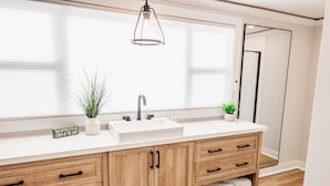 A modern bathroom with a large window, wooden cabinetry, a white sink, and two potted plants. - Lake Winnipesaukee, Weirs Beach, Funspot, Gunstock (Laconia)