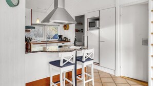 A compact kitchen with a white stove, a microwave, and a sink. There are two white chairs with blue cushions. A green clock is mounted on the wall.