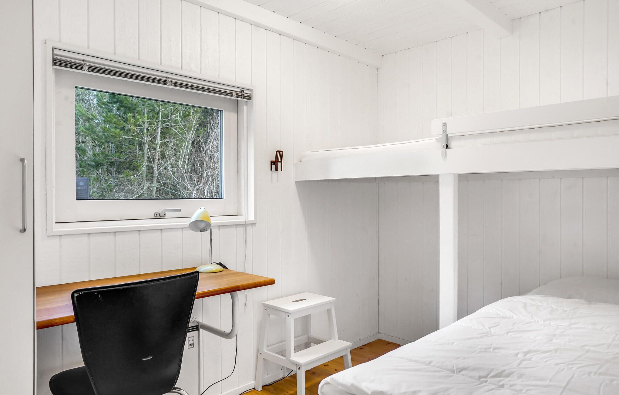 A bedroom with a bunk bed, a desk with a chair, a window with a view of trees, and a bedside lamp.