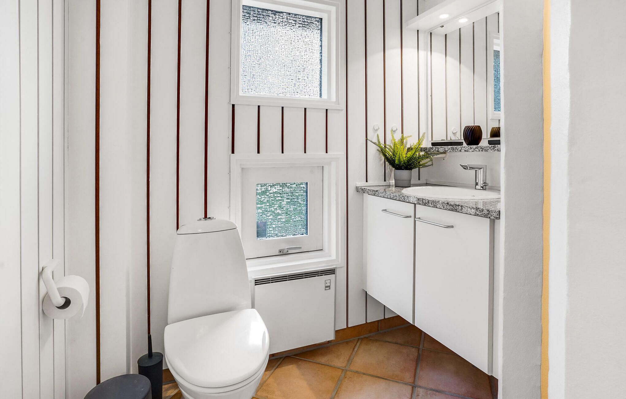 Un baño moderno con inodoro, una ventana con vistas, lavamanos con espejo y una planta en maceta.