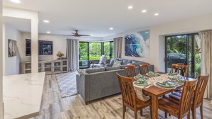A modern living room with a grey sofa, a dining table set for four, and large windows offering a view of greenery. - Stunning 2-bedroom Wailea Elua, walk to beach, ocean view!  (Kihei)