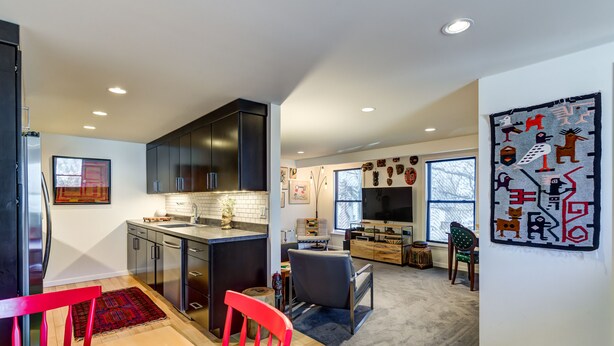 A modern kitchen with dark cabinets, a wooden dining table, and a red chair. There is a TV in the living area with a large window, and a wall with tribal art. - Webster Groves detached Mother-in-Law Suite (Webster Groves)