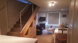 A modern living room with a staircase, a flat-screen TV, a fireplace, and a dining area.