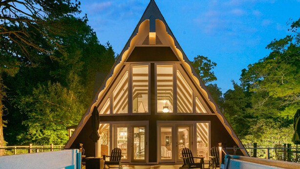 A modern, triangular-shaped building with a large glass facade, a hot tub in the foreground, and a deck area with chairs.