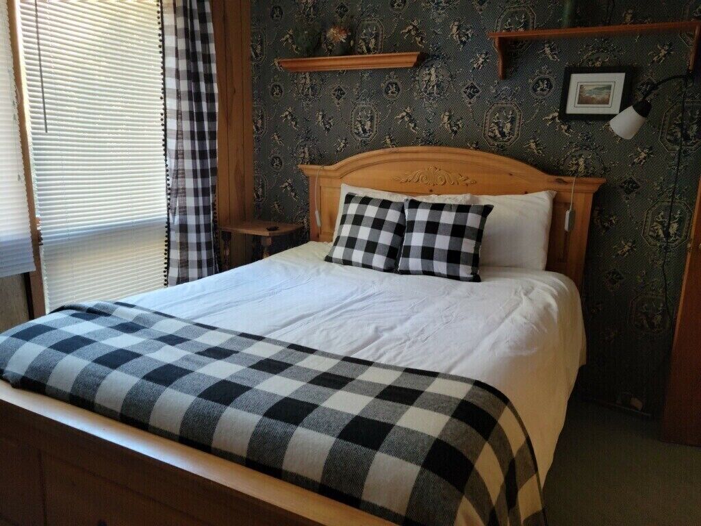 Een netjes opgemaakt bed met een geruit sprei en kussens, een houten hoofdbord, een nachtkastje met een lamp en behang met een patroon.