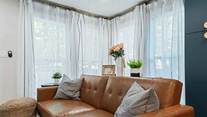 A brown leather sofa with cushions, a small round ottoman, and a vase with flowers on a windowsill. - Cozy Caldwell Retreat | Golf Course Views Near Downtown (Caldwell)