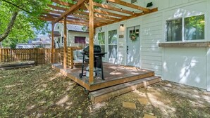 A covered patio with a grill, wooden pergola, and a white house with a window and door. - Cozy Caldwell Retreat | Golf Course Views Near Downtown (Caldwell)