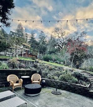 An outdoor patio area with wicker chairs and a round table, surrounded by a garden with various plants and trees. - 🌿5.0 Rating🌿 Sunny, spacious cozy retreat + firepit. World-class trails! (San Geronimo)