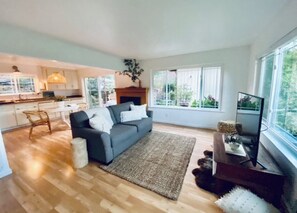 A spacious living area with a grey sofa, a coffee table, and a TV on a wooden stand. There is a large window providing a view of greenery outside. - 🌿5.0 Rating🌿 Sunny, spacious cozy retreat + firepit. World-class trails! (San Geronimo)