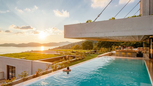 Una piscina con una persona en el agua, rodeada de jardines y una vista del atardecer sobre una masa de agua y colinas.