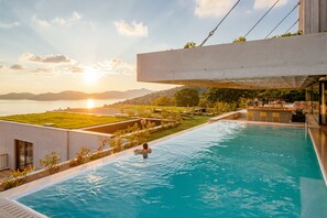 Una piscina con una persona en el agua, rodeada de jardines y una vista del atardecer sobre una masa de agua y colinas.