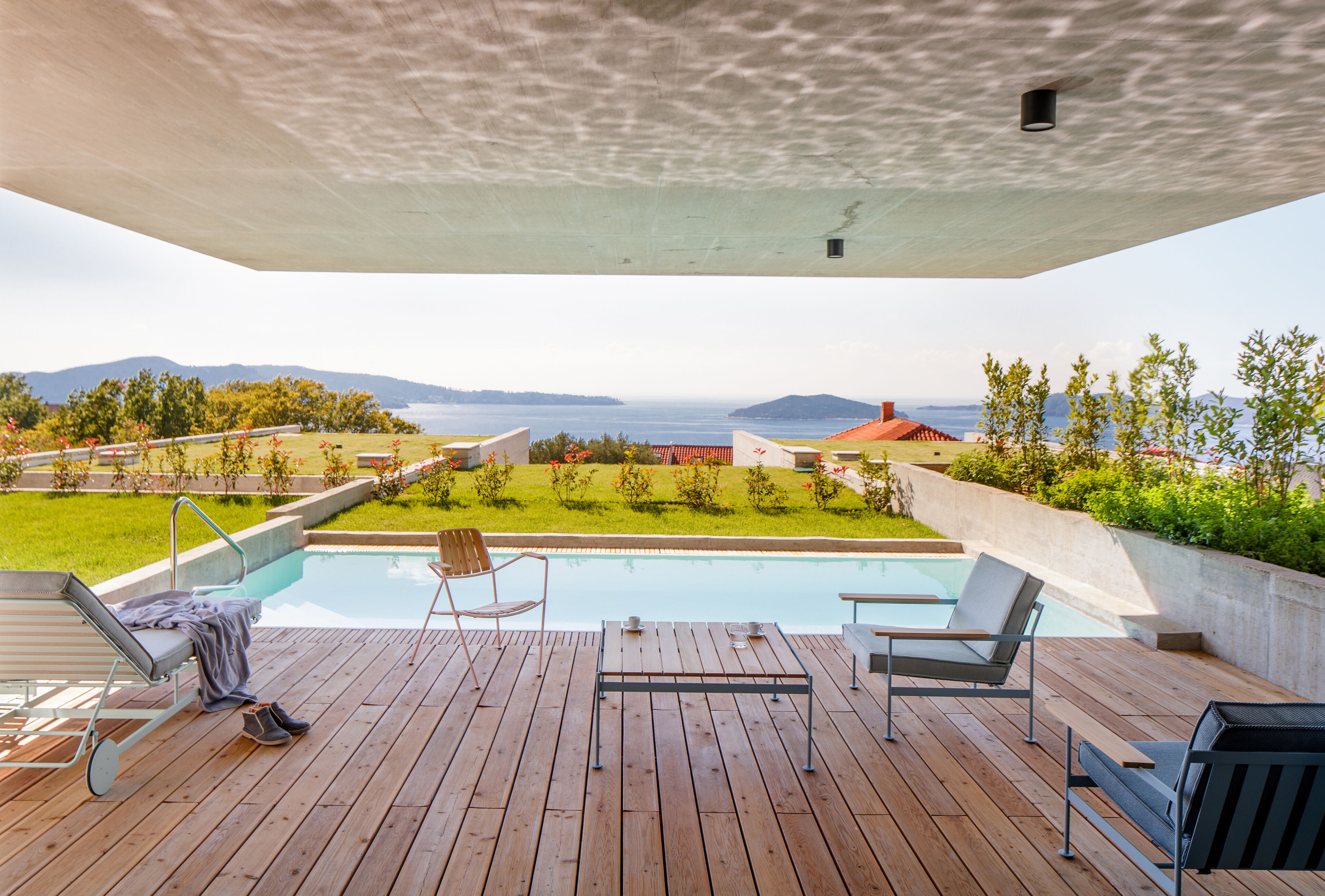 Una terraza de madera con piscina, sillas de descanso y vistas al mar y las colinas.