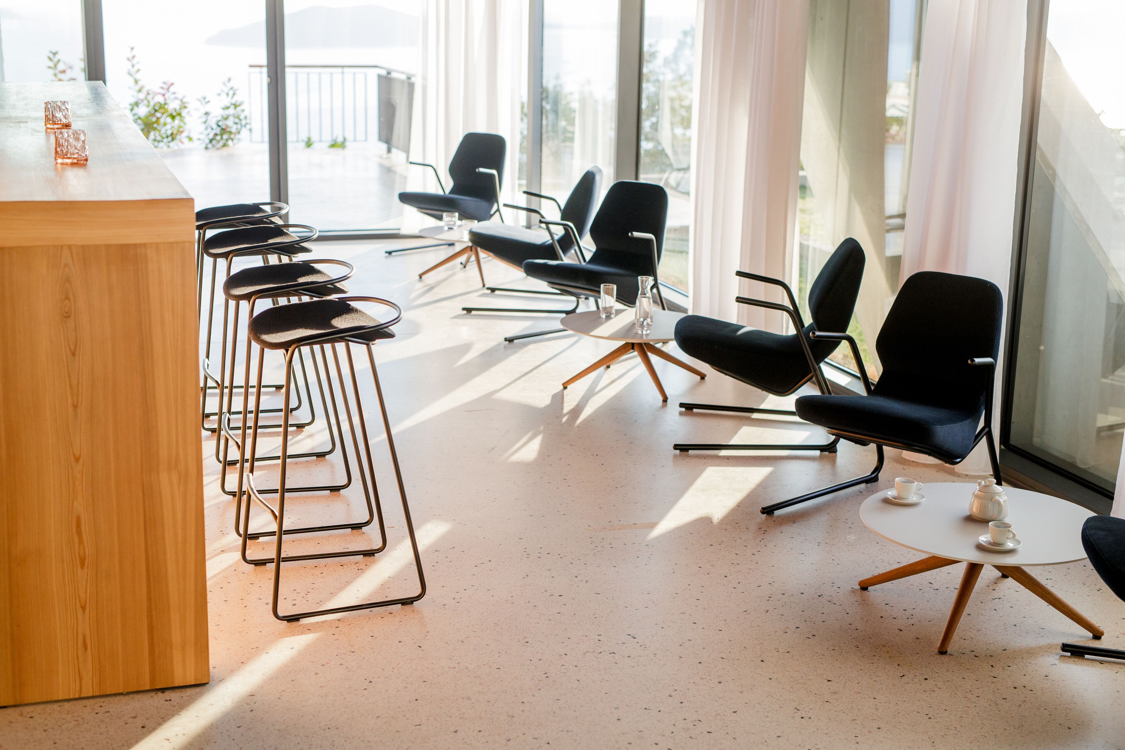 A modern bar area with a wooden counter and black bar stools, arranged in a row with a large window in the background.