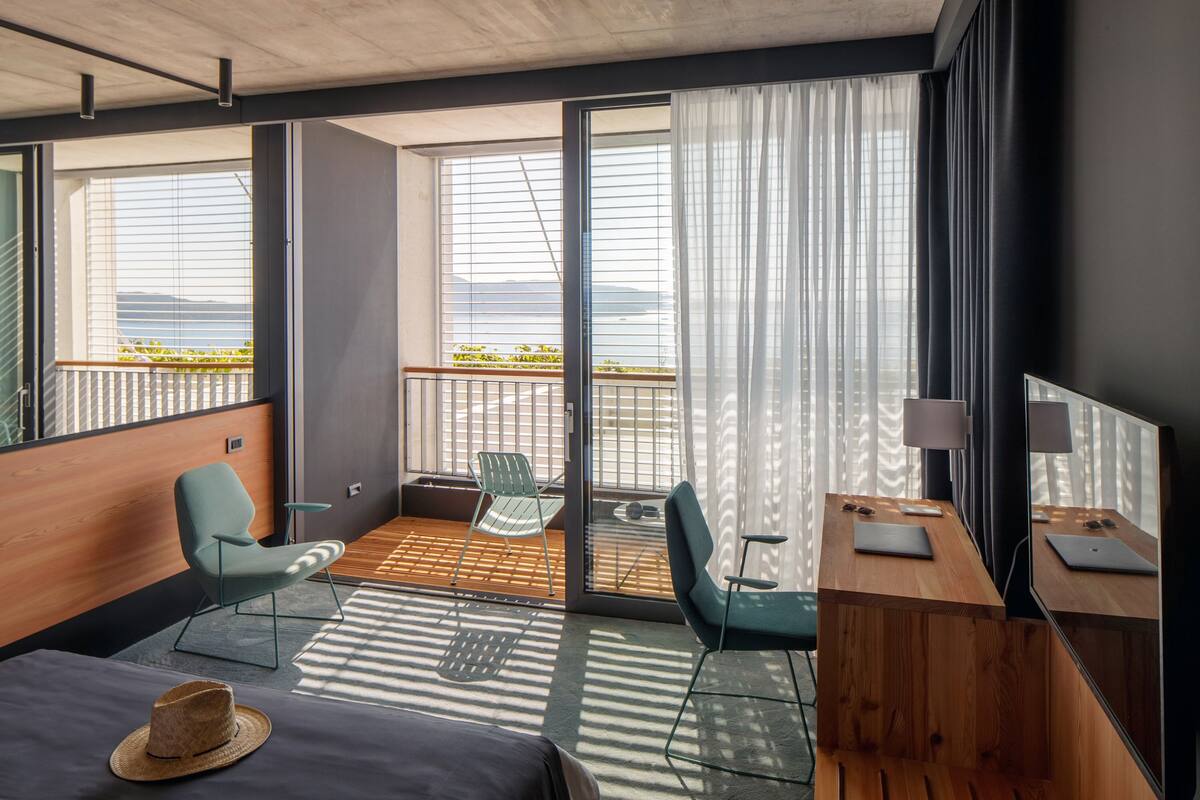 A modern hotel room with a large sliding glass door to a balcony, two chairs, a small table, and a bed with a hat on it.