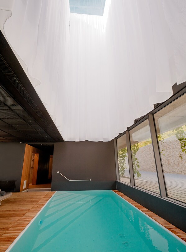 Una piscina interior moderna con tragaluces, suelo de madera y vistas a un muro de piedra y vegetación exterior.