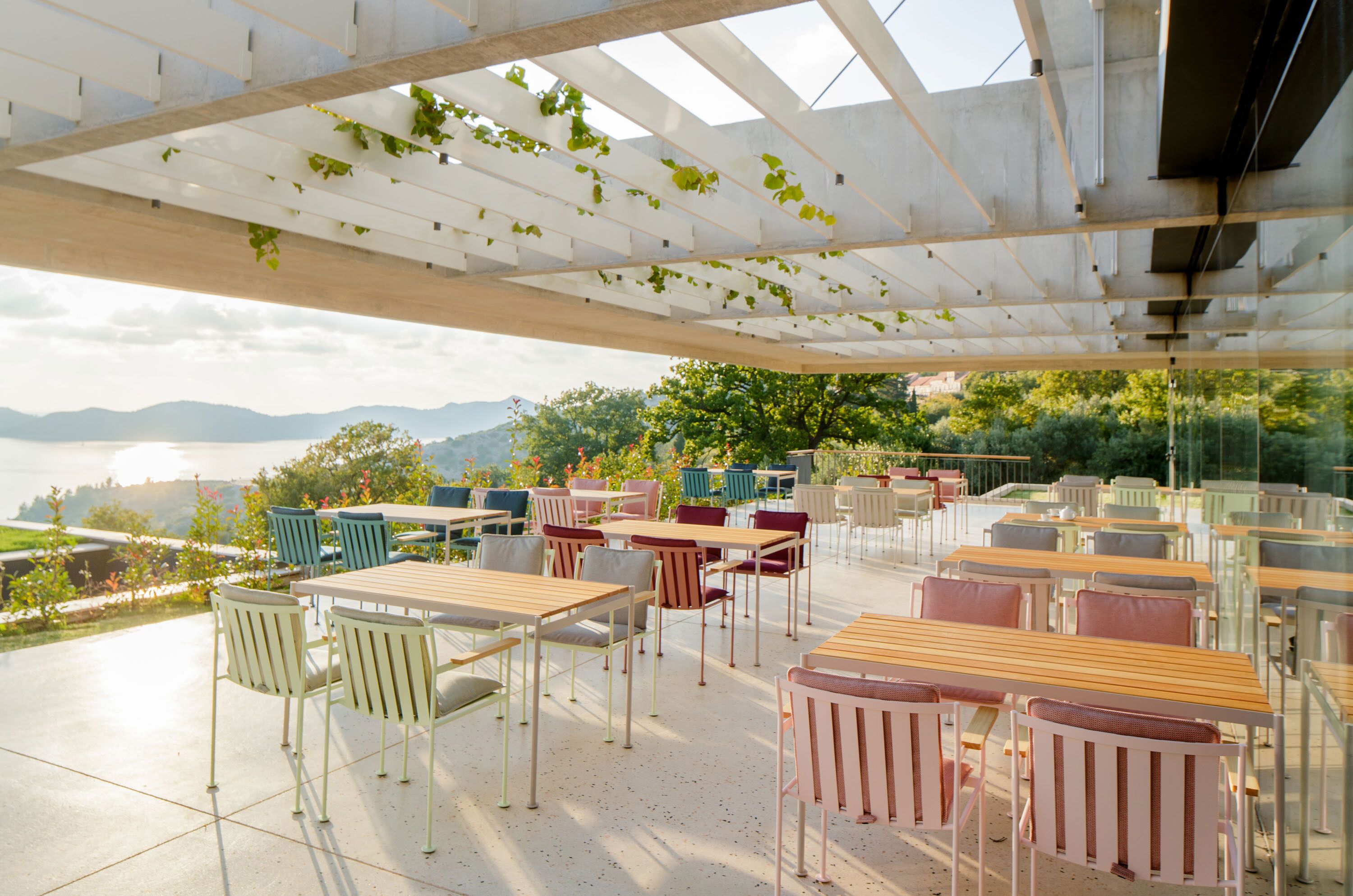 An outdoor seating area with tables and chairs, a pergola with vines, and a view of mountains and greenery.
