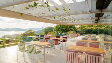 An outdoor seating area with tables and chairs, a pergola with vines, and a view of mountains and greenery.